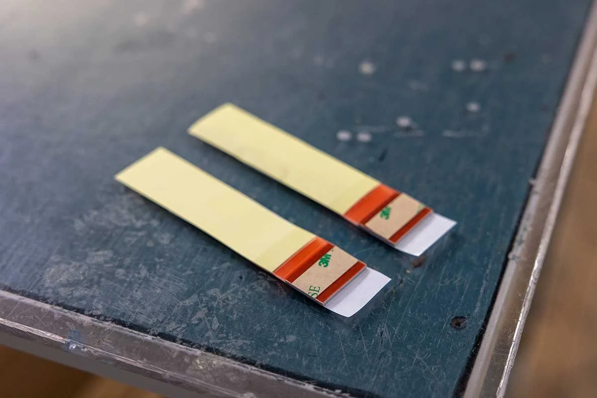 Two adhesive glue strips with orange and green printed backing on a worn green table surface.