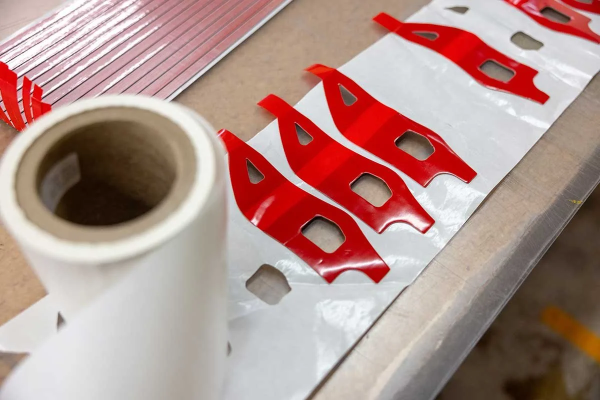 Red and white decal sheets with cut-out letters, a roll of white adhesive film, and a perforated metal sheet on a worktable.