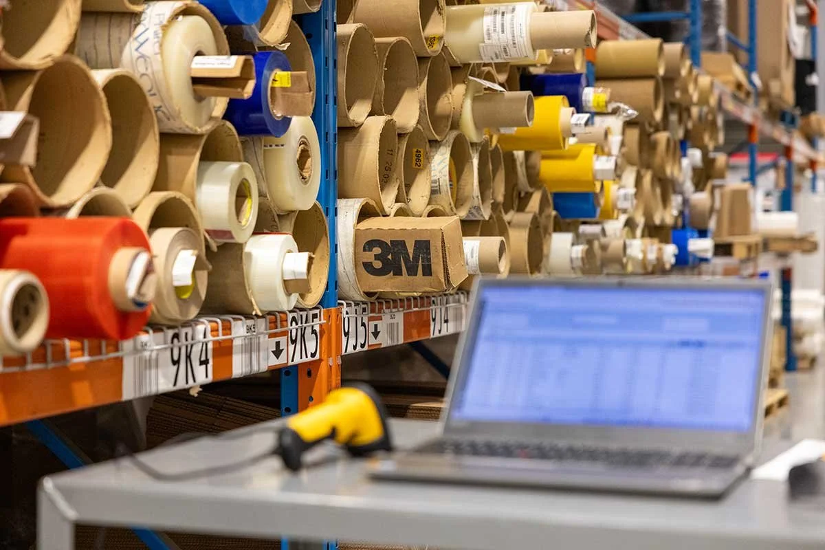 Shelves filled with rolls of tape and a laptop on a work table in a warehouse or hardware store.